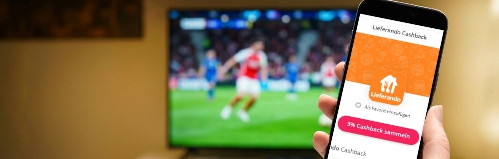 Smartphone mit Lieferando-Seite auf topcashback.de vor einem Fernseher mit laufendem Fußballspiel.
